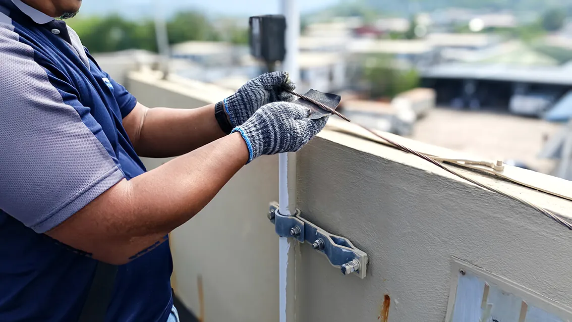 Técnico com luvas fixando um suporte metálico no parapeito da cobertura de uma edificação